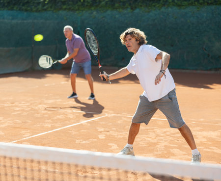 Two men in sportswear playing tennis match on tennis courts, male player receiving hit from tennis racketの写真素材