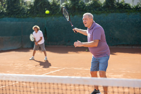 Active aged man hits tennis ball with racket in front of players in court. Two sportive men enjoying doubles matchの写真素材