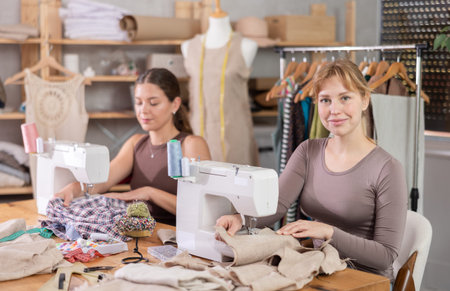 Two positive female dressmakers working with sewing-machine in tailors workshop