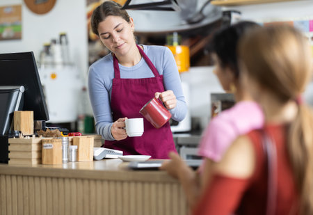 Young female barista pouring coffee into cup at counter while clients waitingの写真素材