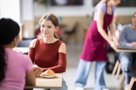 Young girl chatting with friend over coffee and croissant in cafeteriaの写真素材