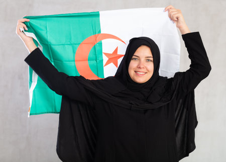 smiling Muslim woman in traditional black hijab holds flag of Algeriaの写真素材