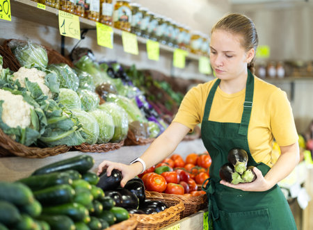 Teenage girl seller puts eggplants in vegetable shopの写真素材