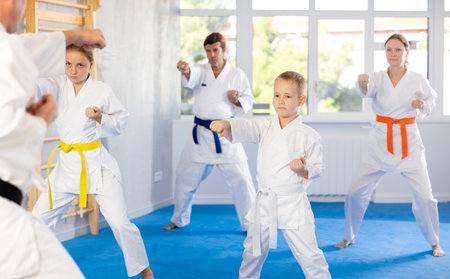 Boy with his family in kimonos and colored belts practicing karate with punches during group martial arts class in gym, accompanied by trainerの写真素材