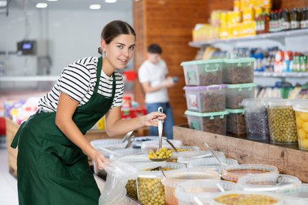 Young girl in supermarket, scooping marinated olives from bucket with ladle