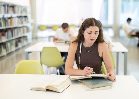 Female student sits at table in library and studies, reads books and takes notesの写真素材
