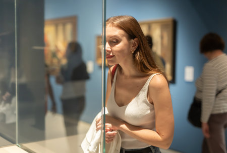 Young female visitor stands near glass display case and examines valuable exhibitの写真素材