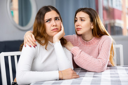 Upset woman sitting at home table with disgruntled woman behind