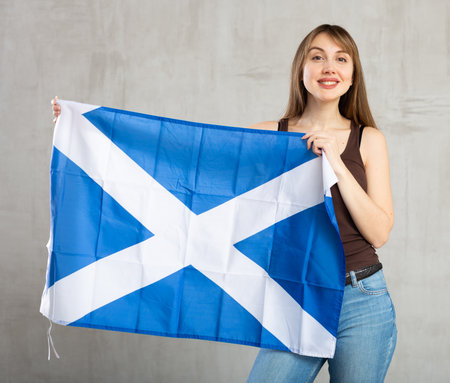 Portrait of confident smiling young female student standing against gray wall with national flag of Scotlandの写真素材