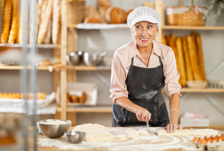 Senior woman works in bakery as baker, cuts dough into portions, form pieces of dough to create bunsの写真素材