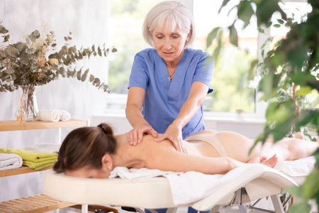 Skilled aged masseuse doing back pain relief massage to young womanの写真素材