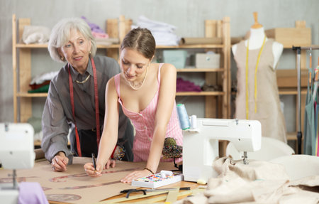 Elderly woman seamstress teaches young woman to draw pattern on paperの写真素材