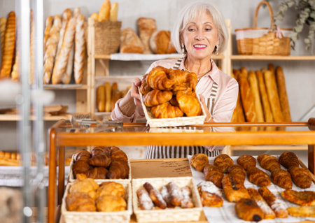 Elderly bakery saleswoman places fresh croissants on displayの写真素材
