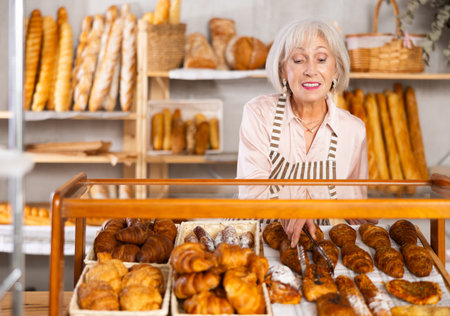 Elderly bakery saleswoman places fresh croissants on displayの写真素材