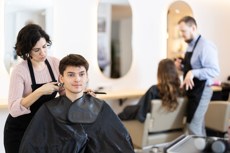 Adult woman hairdresser gives haircut to young guyの写真素材