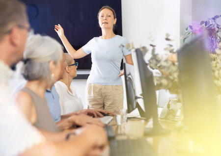 Female IT teacher conducts lesson teaching elderly people on computer in nursing home