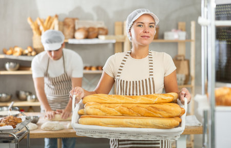 Smiling female baker presenting fresh baguettes in bakeryの写真素材