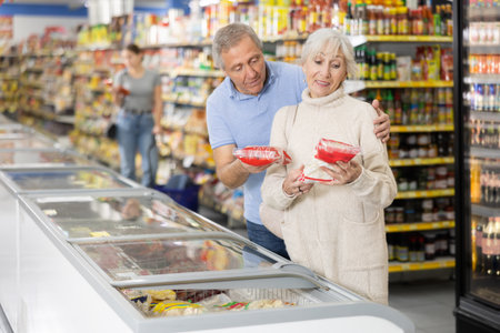 Mature couple shopping in supermarket, choosing frozen convenience food for quick dinnerの写真素材