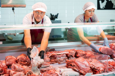 Near counter mature male seller offers shows pork ear in butcher shopの写真素材