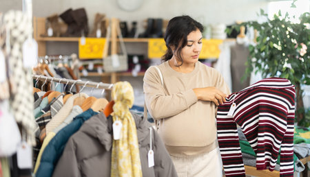 Interested positive pregnant latina young woman shopping sweaters in store. Sale of winter clothing...