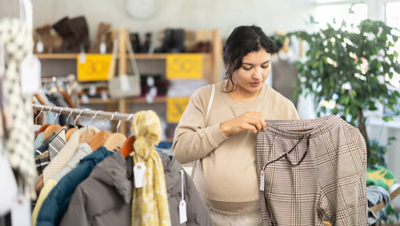 Pregnant woman is busy looking at clothes items in store, she chooses trousersの写真素材