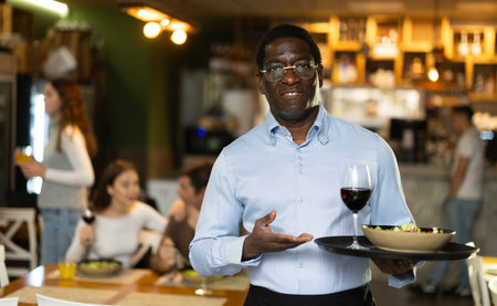 African man of public catering cafe carries order to client, ready to serve guest of cafe establishment.の写真素材
