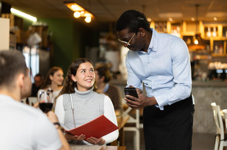 African male waiter communicates with female customer, serve couple in restaurantの写真素材