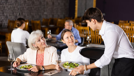 Young male waiter brought food to order of young girl and elderly mother to visitors of restaurant.の写真素材