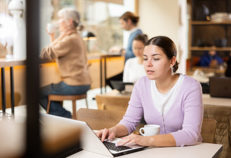 Smiling woman using laptop and drinking coffee in modern cafeの写真素材
