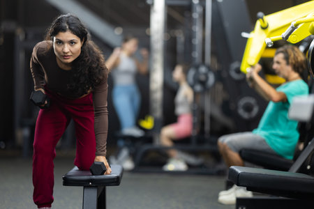Latina woman doing dumbbell rows near bench in hall