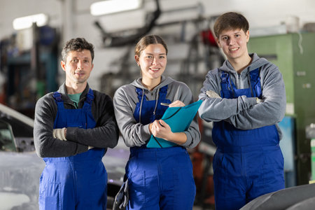 Team of auto mechanics, two men and one woman, wearing blue overalls and posing, ready to provide...
