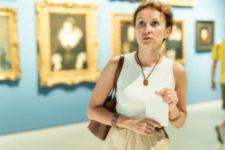 Brunette woman visitor holding guide list standing near pictures in museum of artsの写真素材