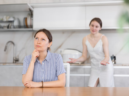 Offended mother sitting at the kitchen table, daughter standing behindの写真素材