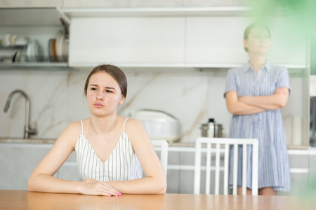 Offended young girl sitting at the kitchen table, angry mother standing behindの写真素材