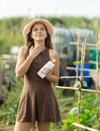 Smiling girl in dress and hat applying sunscreen to skin while gardeningの写真素材