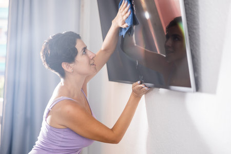 Positive young brunette woman cleaning dust from TV screen