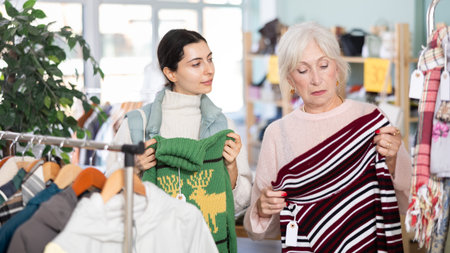 Two focused female clients carefully looking at sweaters for autumn-winter season in clothing store. Family shopping conceptの写真素材