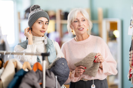 Two happy women choosing warm hat for stylish autumn look in clothing boutiqueの写真素材