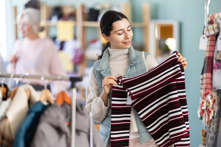 Interested young girl buyer standing in clothing market and carefully examines selection of sweaters on sale of winter clothing collectionの写真素材
