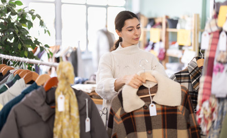 Positive young girl client standing against background of shelves with clothes in modern store and choosing coat or jacketの写真素材