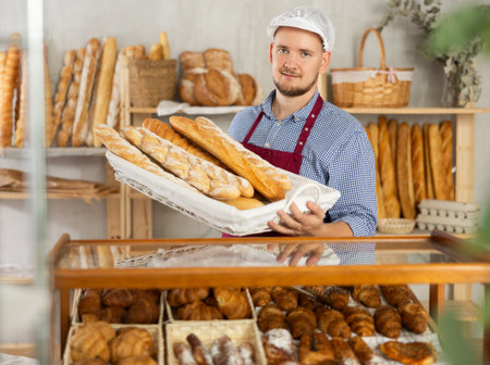 Male seller stands at bakery counter with basket filled by fresh baguettes.の写真素材
