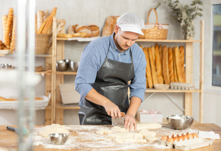 Guy works in bakery as baker, cuts dough into portions, forms pieces of dough to create buns.の写真素材