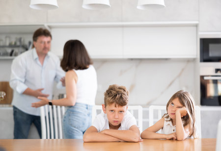 Two children sitting at the kitchen table sadly while their parents quarrellingの写真素材