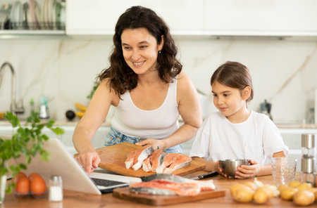 While cooking salmon fillet fish dish, woman and little daughter look at laptopの写真素材