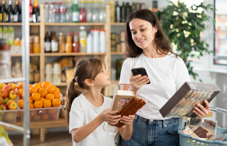 Daughter and mother choose delicious cookies in supermarket - scan QR code on label with smartphoneの写真素材