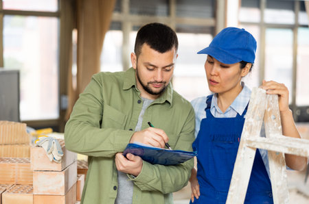 Young designer with papers talking to female builder in apartment during renovation