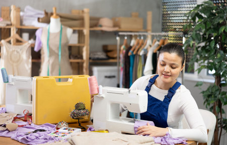 Positive young female dressmaker working with sewing-machine in tailors workshop