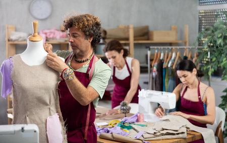 Male tailor sews new dress on mannequin in a sewing studio. Other woman tailor is sewing on sewing...