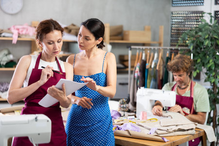 Two female dressmakers discussing dress sketches on paper. Man works on sewing machine.の写真素材