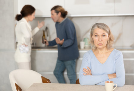 Senior woman is sitting in kitchen during quarrel between her son and his wifeの写真素材